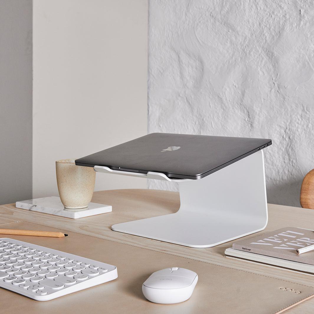 Werkplek op licht eiken tafel met een gesloten laptop op een witte laptop standaard met bijpassende bureau accessoires in wit en sand kleur onderlegger