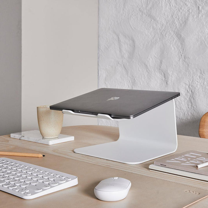 Werkplek op licht eiken tafel met een gesloten laptop op een witte laptop standaard met bijpassende bureau accessoires in wit en sand kleur onderlegger
