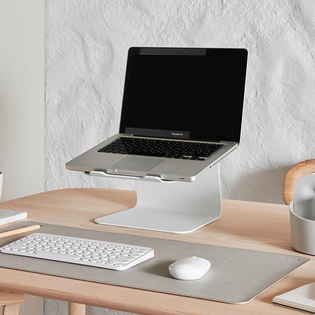 Stijlvolle werkplek met een laptop op witte standaard en een wit toetsenbord en muis op een stone kleurige bureau onderlegger