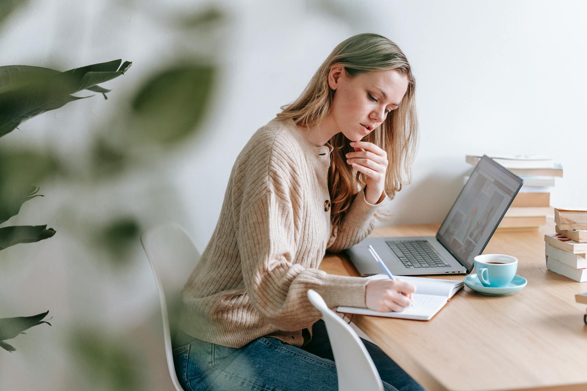 Vrouw is thuis aan het werk aan tafel achter haar laptop en maakt een aantekening in notitieboekje